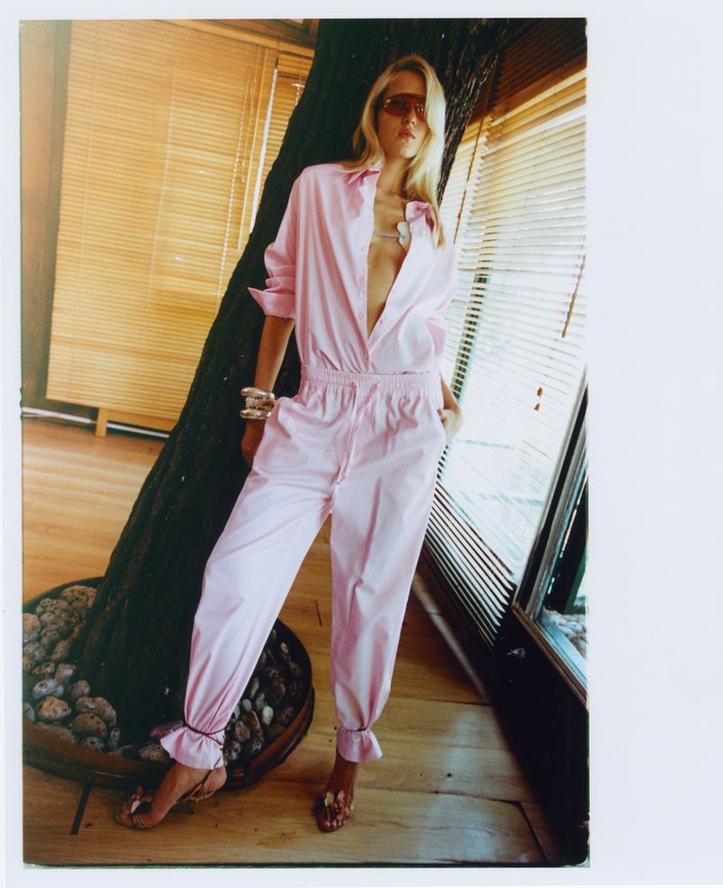 A woman in a pink jumpsuit poses indoors with light coming through blinds. She is wearing sunglasses and gold bracelets.