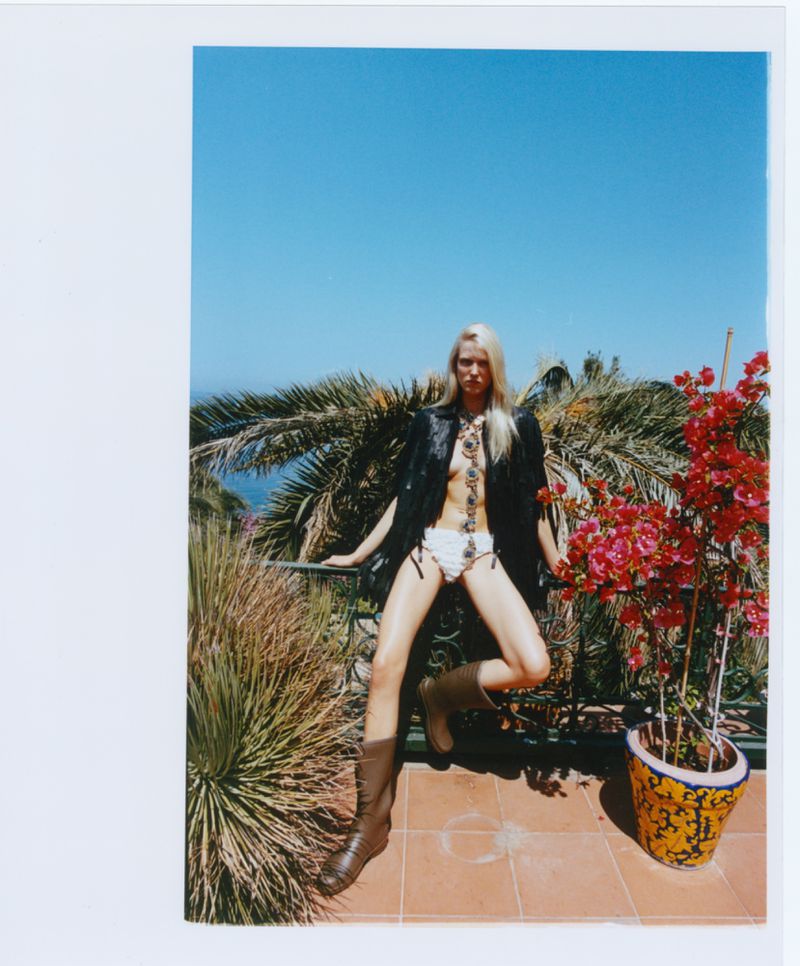 A blonde fashion model in a black jacket, white bottoms, and brown boots poses on a sunlit balcony with palm trees and red flowers.