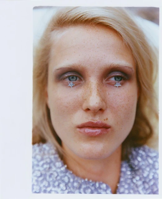 Close-up portrait of a blonde woman wearing star-shaped makeup around her eyes and a light purple sequined top.
