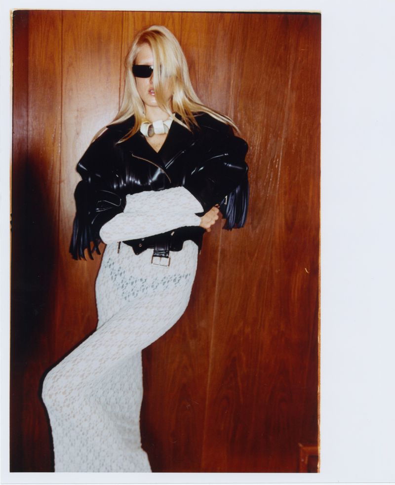 Fashion photo of a blonde woman posing in a black leather jacket and white skirt against a wood panel wall, wearing sunglasses.