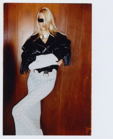 Fashion photo of a blonde woman posing in a black leather jacket and white skirt against a wood panel wall, wearing sunglasses.