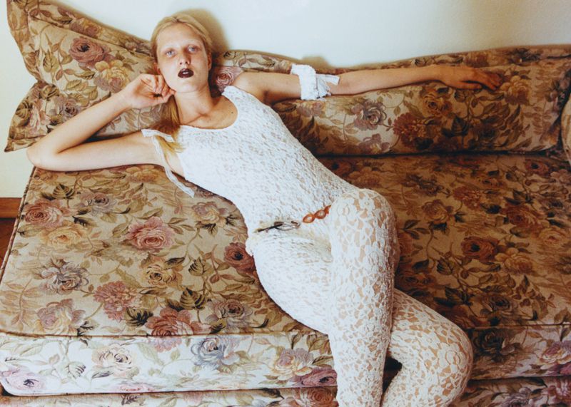 Fashion photo of a fair-skinned adult woman lying on a floral couch. She wears a white lace bodysuit with dark lipstick and neutral background.