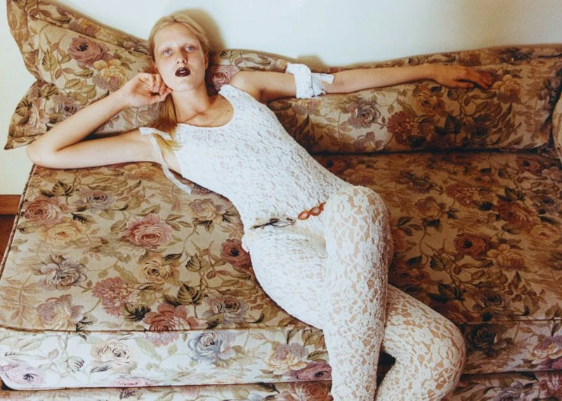 Fashion photo of a fair-skinned adult woman lying on a floral couch. She wears a white lace bodysuit with dark lipstick and neutral background.