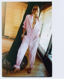 Model Vakare Steputyte in a pastel pink unbuttoned jumpsuit and tinted sunglasses leaning against an indoor tree by window blinds.