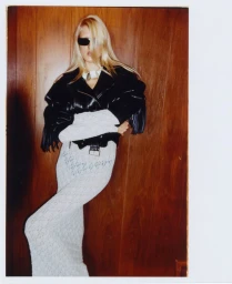 Model wearing dark sunglasses, a black fringe leather jacket, and a white lace dress against a wood panel wall