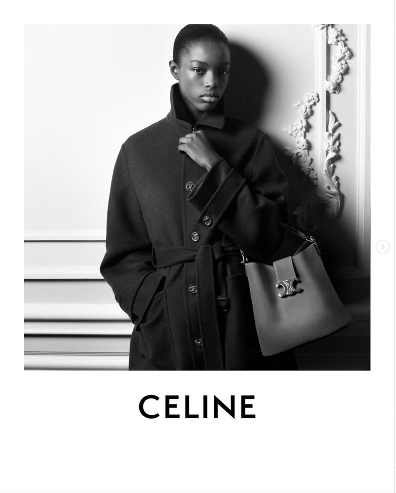 Black and white fashion shot of a woman wearing a dark coat and holding a structured leather handbag in an indoor setting.