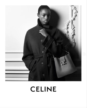 Black and white fashion shot of a woman wearing a dark coat and holding a structured leather handbag in an indoor setting.