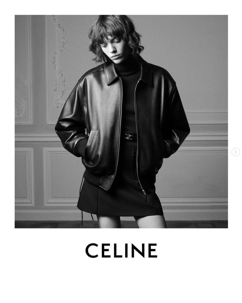 Black and white fashion shot of a female model wearing a leather jacket and short skirt against a decorative wall. Studio lighting.