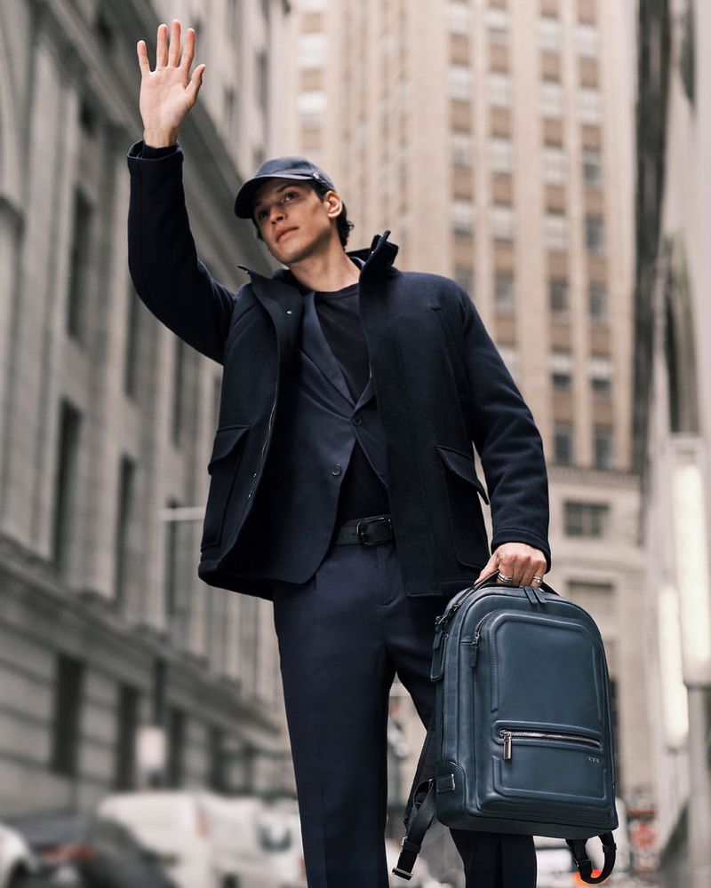 Man in a suit and cap waves while holding a backpack in a city environment with tall buildings in the background, daytime.