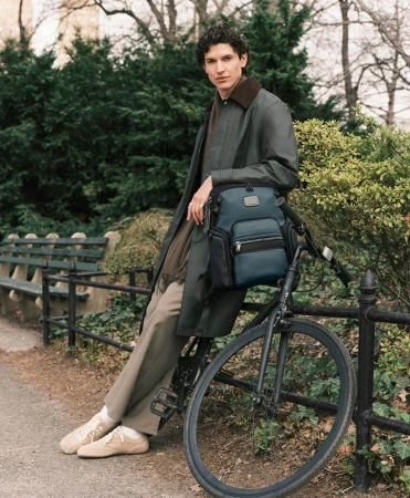 Model Kyle Vieira in a long green coat leaning against a bicycle holding a blue Tumi backpack along a park path.