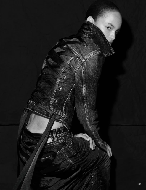 A model in a dark denim jacket with lace-up back and matching jeans, looking over shoulder in a studio portrait against a dark backdrop.