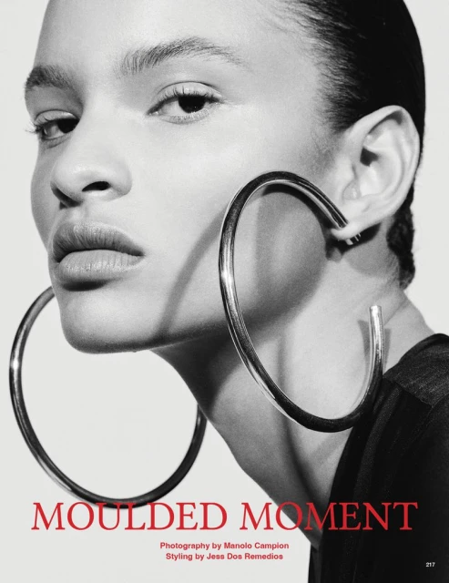 Close-up of a model wearing hoop earrings. The shot is monochrome, highlighting the earring's design and the model's facial features.