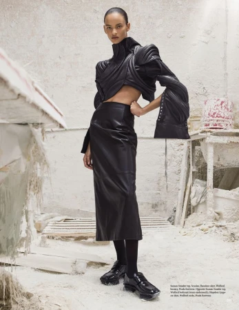 Fashion editorial shot of a woman posing in a black, structured outfit against a textured white backdrop in a photo studio setting.