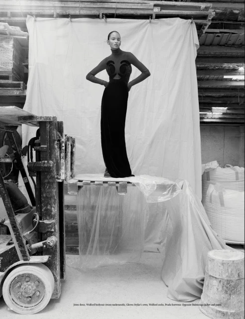 Fashion shot of a woman in a black dress standing on a pallet in a warehouse setting, next to a forklift and under a white sheet.