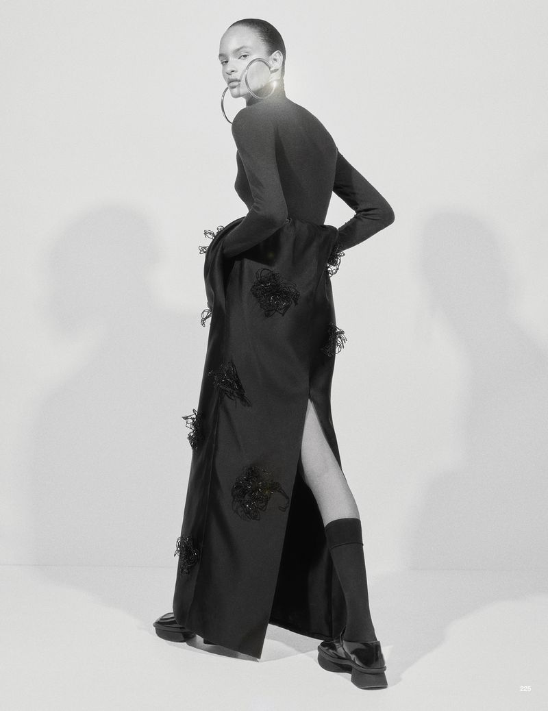 Model Ana Portela wearing a long black embellished gown, oversized hoop earrings, and platform shoes against a studio backdrop.