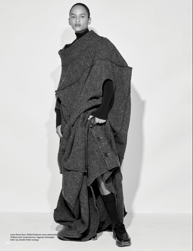 Fashion shot of a young adult wearing a large, grey, wool, avant-garde coat, black turtleneck, and black socks, posing in a studio.