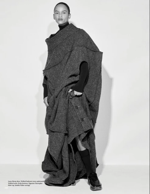 Fashion shot of a young adult wearing a large, grey, wool, avant-garde coat, black turtleneck, and black socks, posing in a studio.