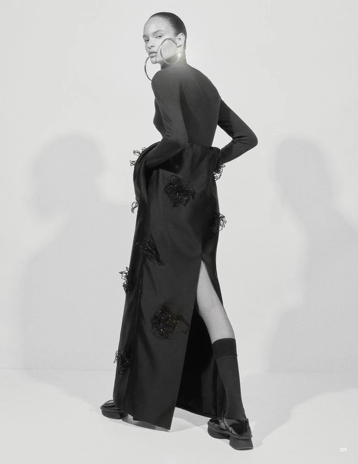 Model Ana Portela wearing a long black embellished gown, oversized hoop earrings, and platform shoes against a studio backdrop.