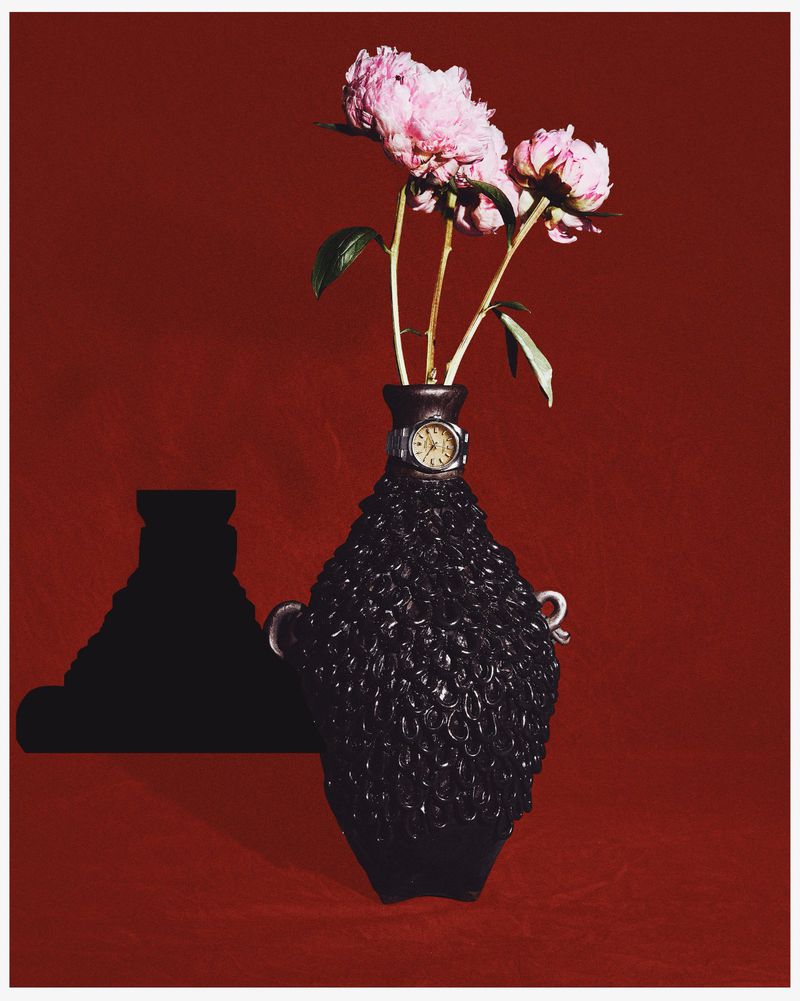 Still life photograph of pink peonies in a black textured vase with a watch, set against a solid deep red background, with dramatic shadows.