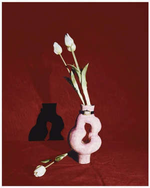 Sculptural pink ceramic vase holding white tulips wrapped with a wristwatch against a textured burgundy fabric backdrop.