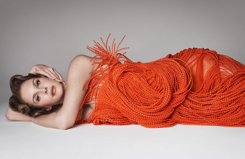 Adult female model poses in a photo studio wearing a textured orange dress and light makeup while lying down against a gray background.