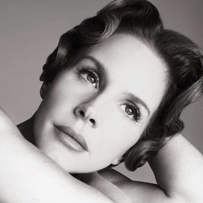 Black-and-white portrait of a fair-skinned adult female with wavy dark hair, makeup, and bare shoulders against a gray background.