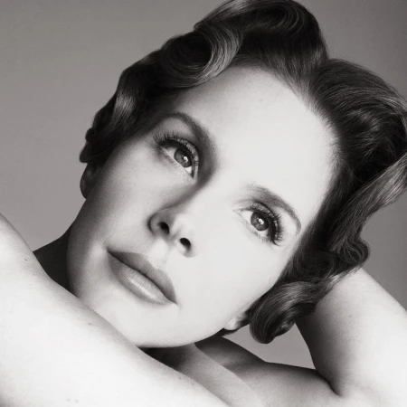Black-and-white portrait of a fair-skinned adult female with wavy dark hair, makeup, and bare shoulders against a gray background.