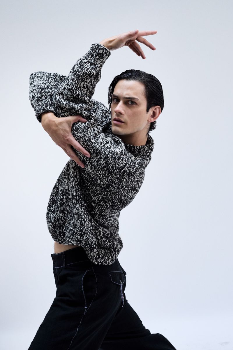 Ballet dancer in a black and white sweater and dark pants strikes a pose in a photo studio against a plain white background.