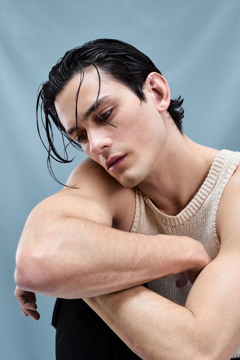 Portrait of a dark-haired adult male wearing a tank top, resting his arms on his knees, with a pale blue background in a studio setting.