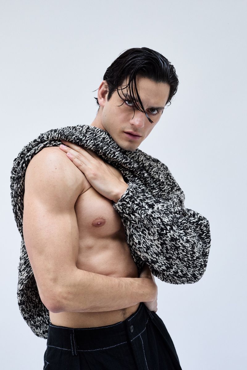 Portrait of a muscular adult male in a black and white sweater, posing shirtless against a white backdrop, with dark hair and intense gaze.