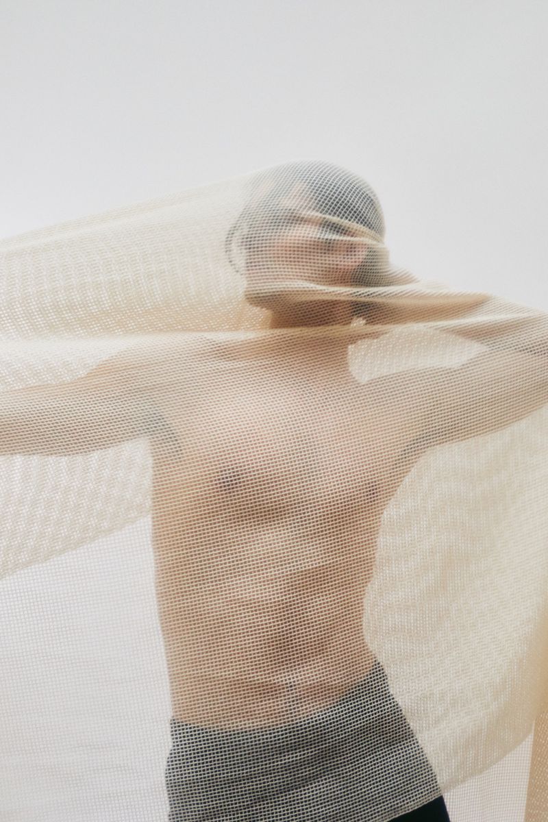 A model stands against a plain backdrop partially obscured by a light beige net-like fabric in a studio setting.