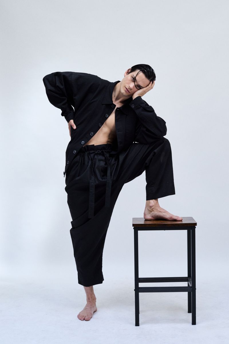 Barefoot male model in an unbuttoned black shirt and oversized trousers resting one foot on a wooden stool against a white backdrop.