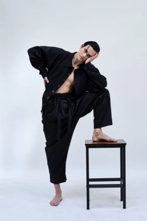 Barefoot male model in an unbuttoned black shirt and oversized trousers resting one foot on a wooden stool against a white backdrop.
