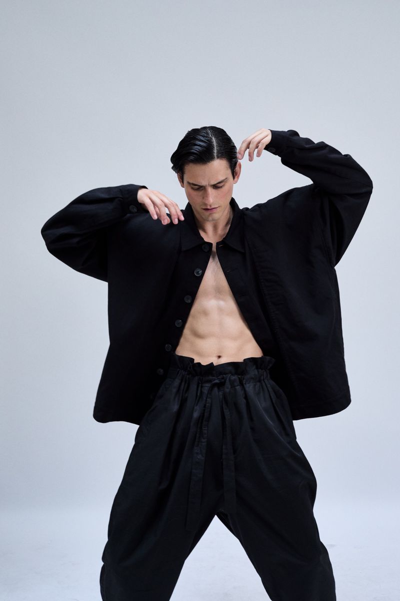Fashion shot of a young adult male model in an unbuttoned black shirt and black pants posing in a photo studio on a white background.