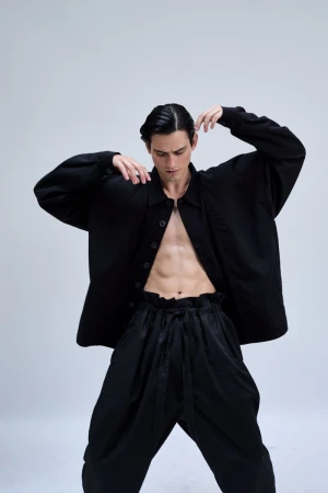 Fashion shot of a young adult male model in an unbuttoned black shirt and black pants posing in a photo studio on a white background.