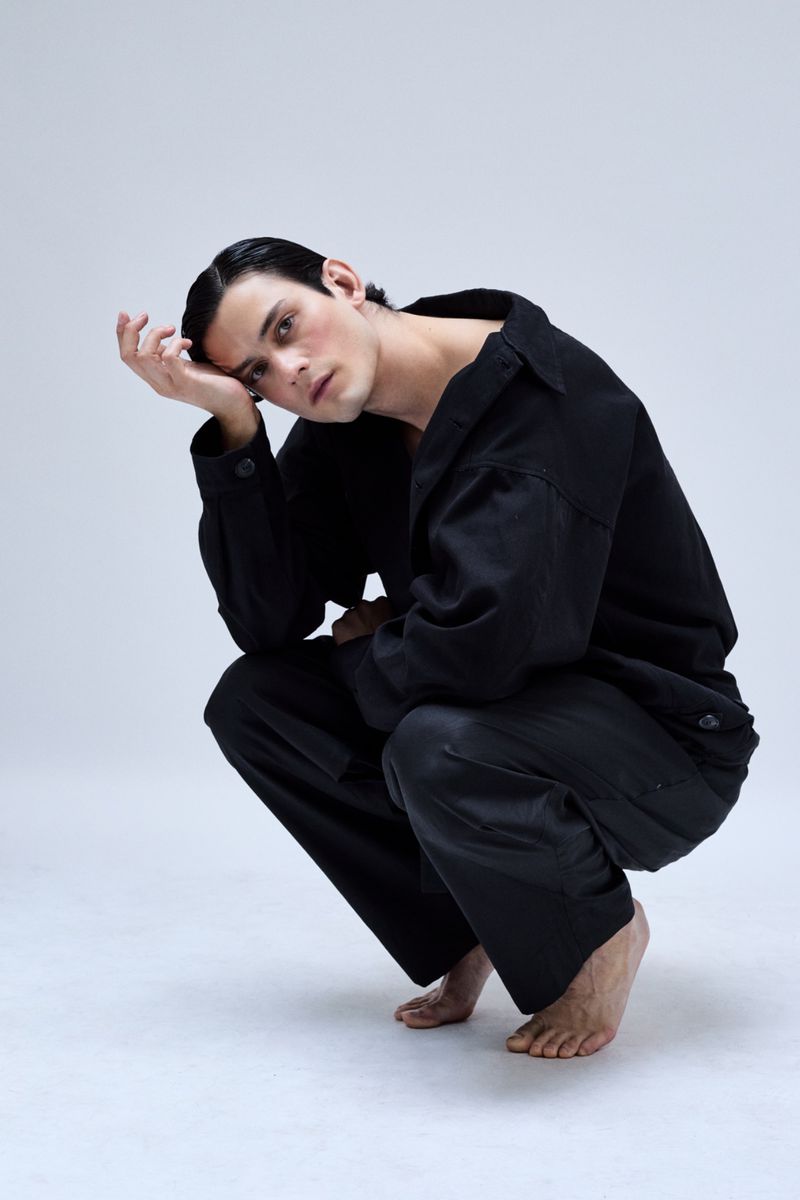 Young adult crouches barefoot in a photo studio, resting head on hand, wearing a black collared shirt and loose pants against a white backdrop.