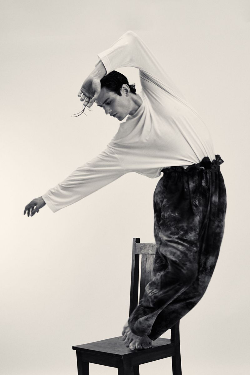 Monochrome portrait of a young adult male balancing barefoot on a wooden chair in a studio, wearing a long-sleeved shirt and tie-dye pants.
