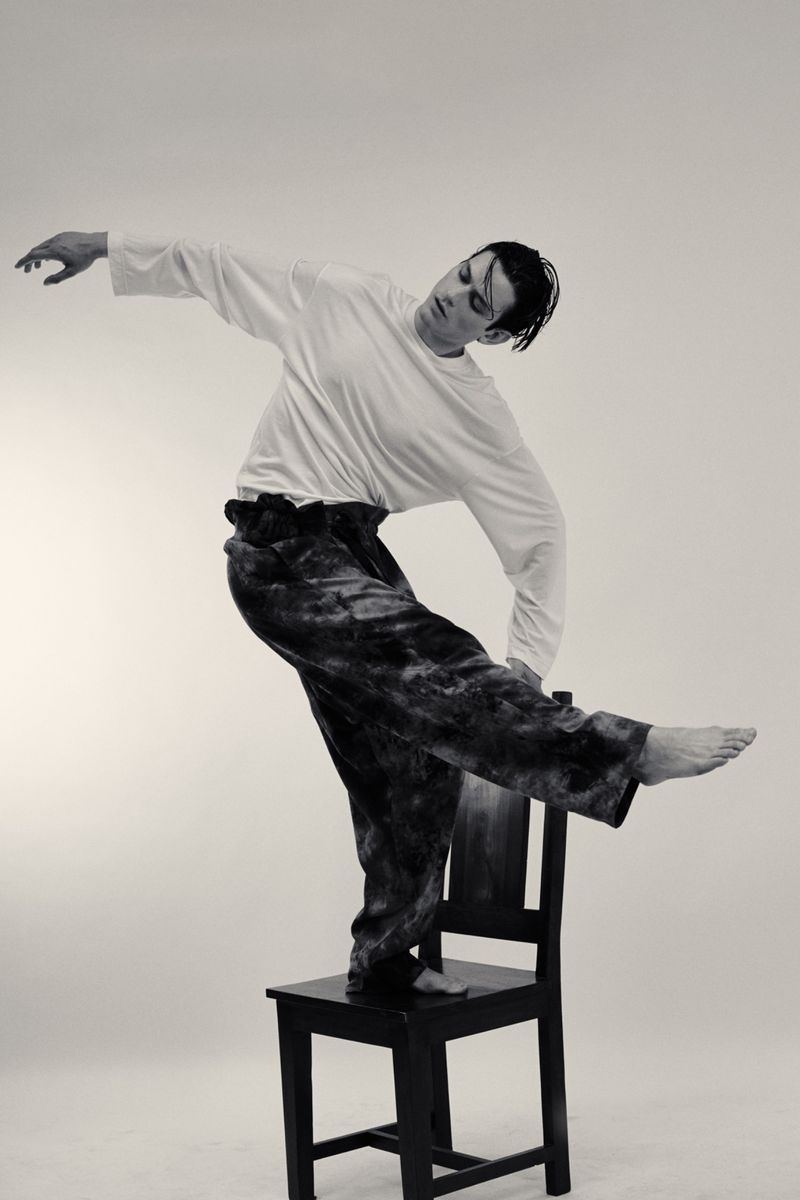Model Tristan Ridel balancing barefoot on a black wooden chair in a white long-sleeve shirt and wide-leg trousers.