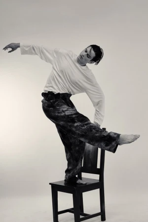 Model Tristan Ridel balancing barefoot on a black wooden chair in a white long-sleeve shirt and wide-leg trousers.