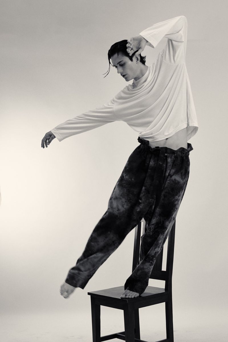 Black and white studio shot of a slim male dancer in tie-dye pants balancing on a chair with outstretched arms and bare feet.