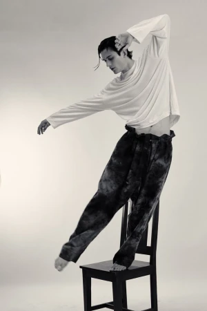 Black and white studio shot of a slim male dancer in tie-dye pants balancing on a chair with outstretched arms and bare feet.