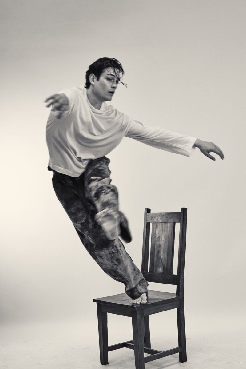 Black and white studio shot of a young adult man balancing on a wooden chair in a white long sleeve t-shirt and patterned pants.