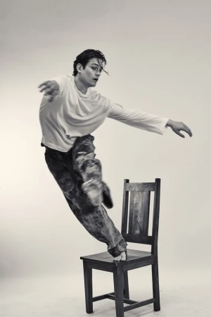 Black and white studio shot of a young adult man balancing on a wooden chair in a white long sleeve t-shirt and patterned pants.