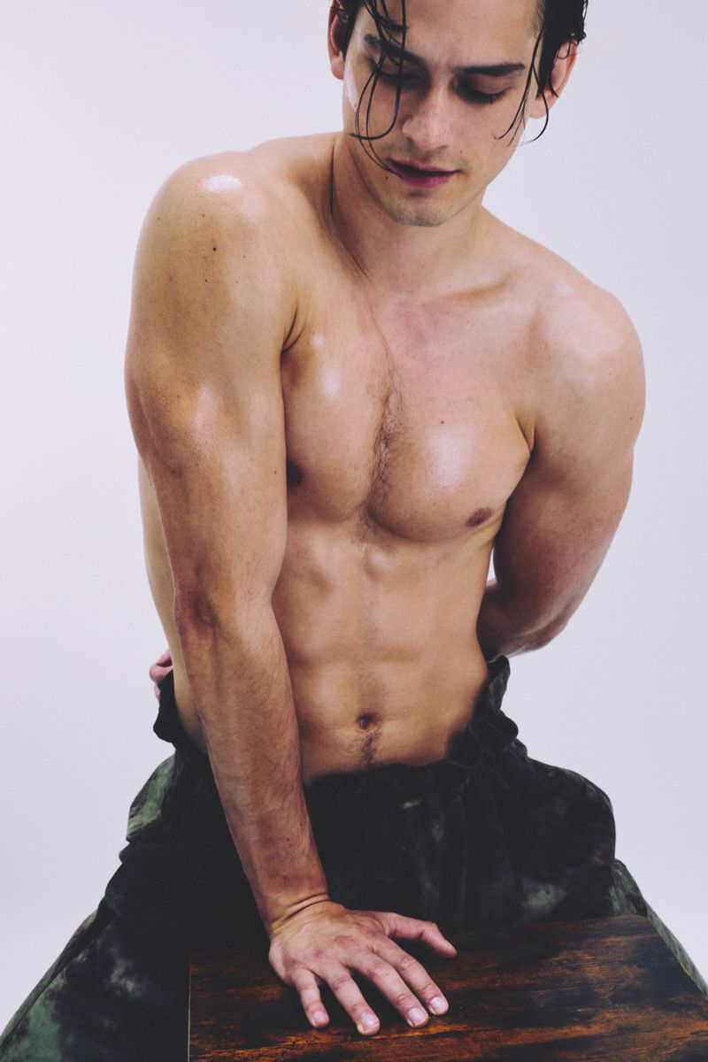Adult male poses shirtless, seated on a wooden block, with wet hair, dark pants, and an athletic build in a bright photo studio.
