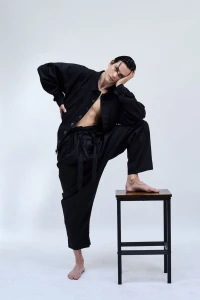 Barefoot male model in an unbuttoned black shirt and oversized trousers resting one foot on a wooden stool against a white backdrop.