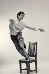 Man in a white long-sleeve shirt and patterned trousers balancing dynamically on a wooden chair against a plain studio background.
