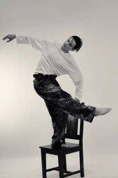 Model Tristan Ridel balancing barefoot on a black wooden chair in a white long-sleeve shirt and wide-leg trousers.