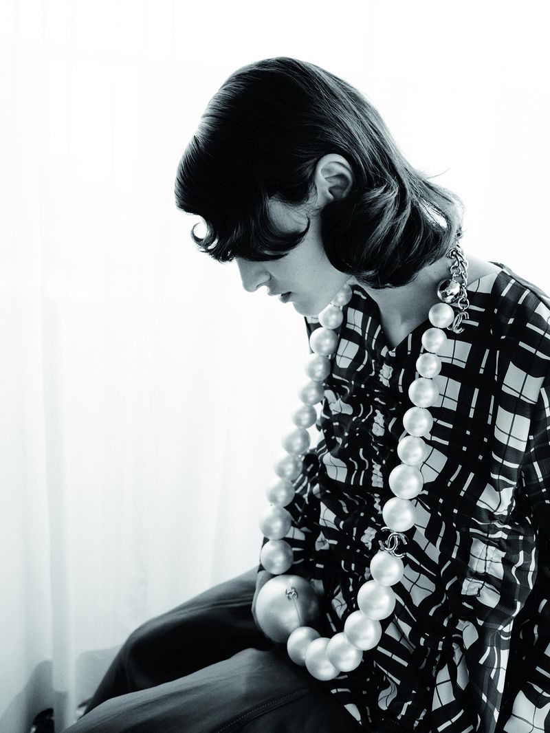 Black and white portrait of a woman wearing a patterned top and a large pearl necklace, looking down with soft window lighting.