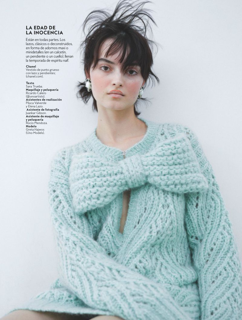 Close-up of a woman with dark hair wearing a light-blue knit sweater with a large bow, posing in front of a white background.