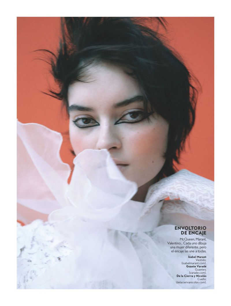 Close-up portrait of a woman with dark hair and dramatic eyeliner, wearing a white ruffled top against an orange backdrop.